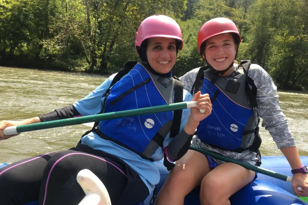 Two people in a raft smiling, both wearing blue lifejackets with a river and greenery behind them.